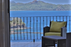 Ein Balkon mit Korbstuhl und Kissen, der Blick auf die Küste mit klarem, blauem Wasser und einer entfernten Insel gewährt.