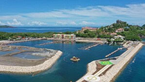 Vue aérienne d’une marina côtière avec de nombreux quais, un grand bâtiment aux façades colorées et un paysage vallonné en arrière-plan.
