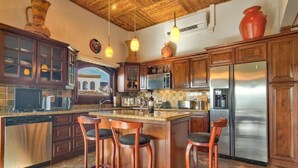 A kitchen with wooden cabinets, a stainless steel refrigerator, and a bar area with stools. - Flamingo Rose Vista in Playa Flamingo (Cabo Velas)