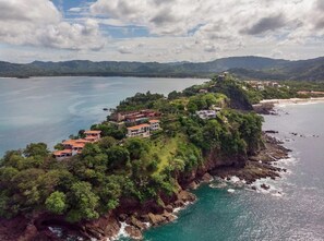 A coastal property with multiple buildings, lush greenery, and a view of the sea and mountains.