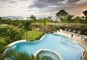 A swimming pool with a curved edge, surrounded by lush greenery and a view of the sea. - Playa Potrero 4 BR Home Large Saltwater Pool Spectacular Views - Villa Oasis (Tempate)