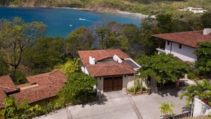 A residential area with houses featuring red-tiled roofs, surrounded by lush greenery and a coastal view.