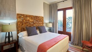 A hotel room with a large bed, two bedside lamps, a wooden headboard, a balcony with curtains, and a chair.