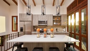 A modern kitchen with a large island, wooden beams, stainless steel appliances, and a large window with sunlight.