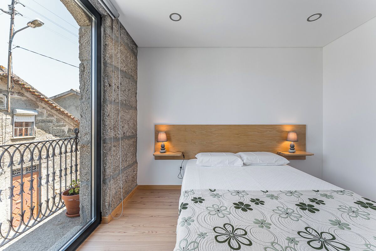 A bedroom with a wooden headboard, a bed with a floral bedspread, two bedside lamps, a potted plant, and a view of a stone building outside.