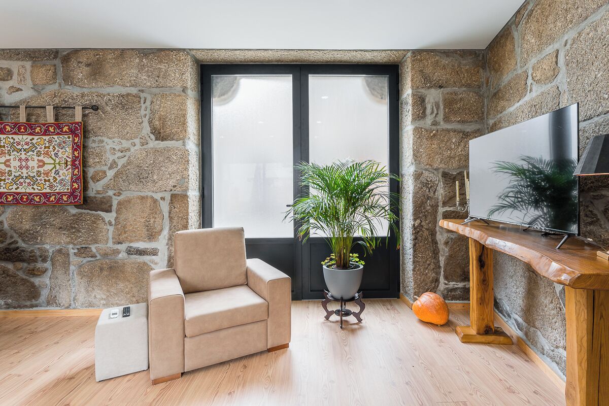 Un salon chaleureux avec un mur en pierre, un canapé beige, une plante en pot, un meuble TV en bois et une grande fenêtre.