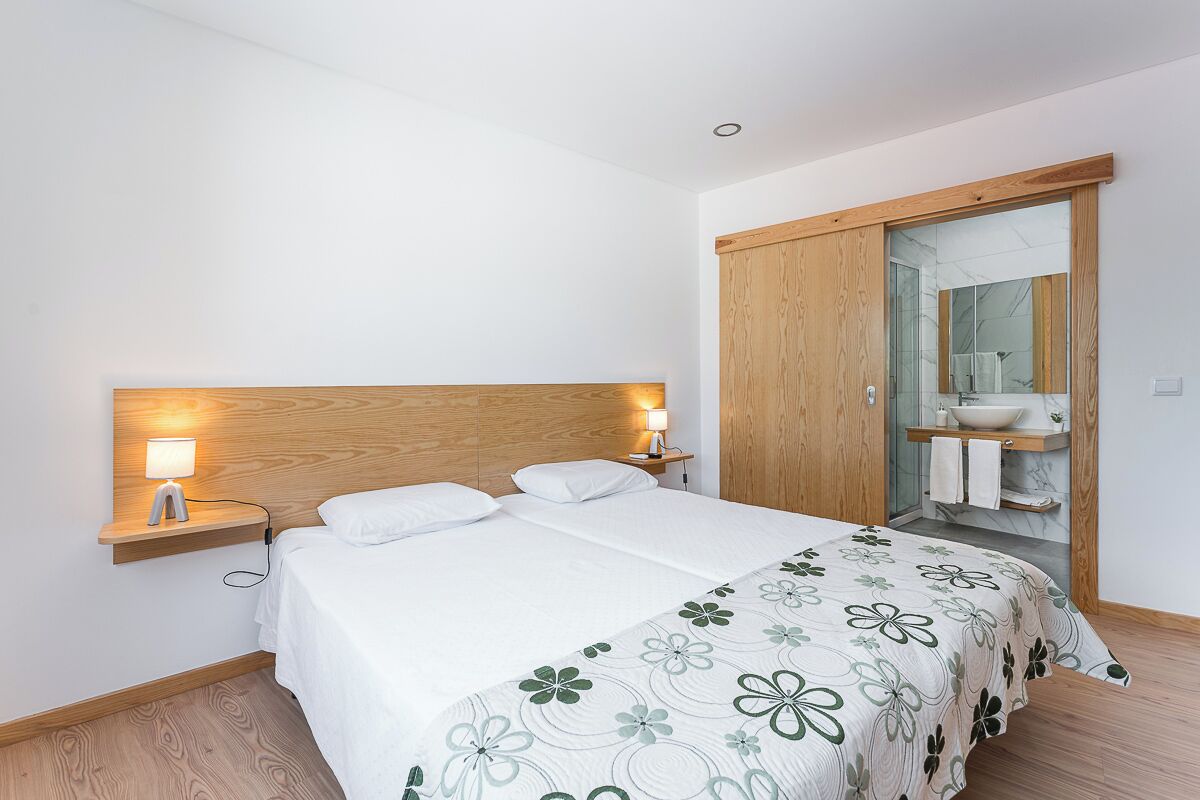A bedroom with a wooden headboard, two bedside tables with lamps, a bed with a floral bedspread, and a sliding door leading to a bathroom with a mirror and sink.