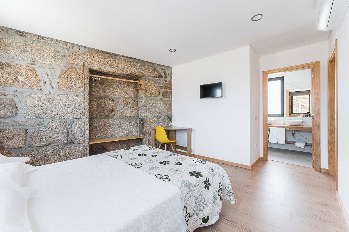 Une chambre avec un mur en pierre, un lit recouvert d’une couvre-lit à motifs floraux, un bureau, une chaise et un téléviseur fixé au mur.