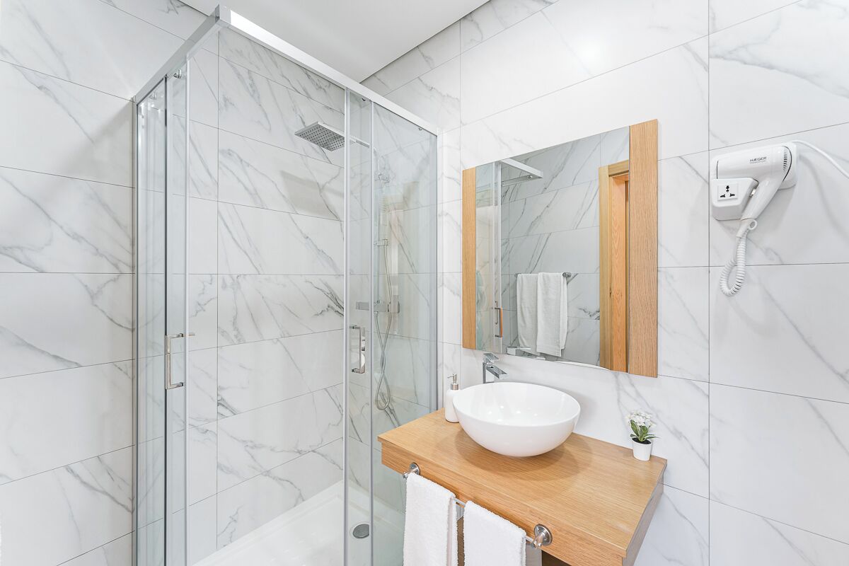 Uma casa de banho moderna com cabine de duche em vidro, toucador de madeira com lavatório branco e secador de cabelo fixado na parede.