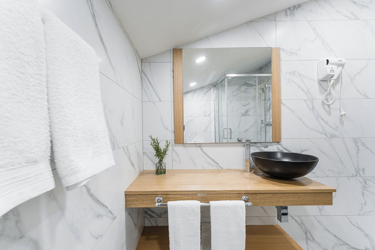 A modern bathroom with a marble wall, a wooden vanity, a black vessel sink, and a handheld hair dryer mounted on the wall.