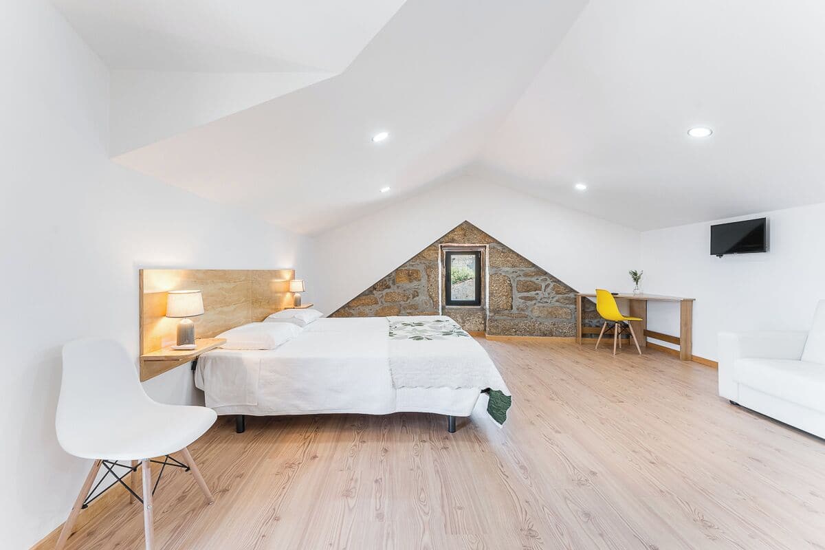Une chambre avec un plafond en pente, un lit, un bureau, une chaise et un téléviseur fixé au mur.