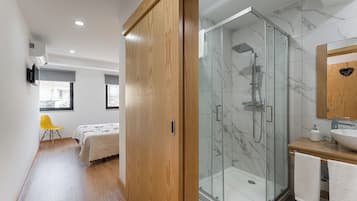 Ein modernes Badezimmer mit Duschkabine aus Glas, einem Holzkosmetikschrank und einem Bett im angrenzenden Zimmer.