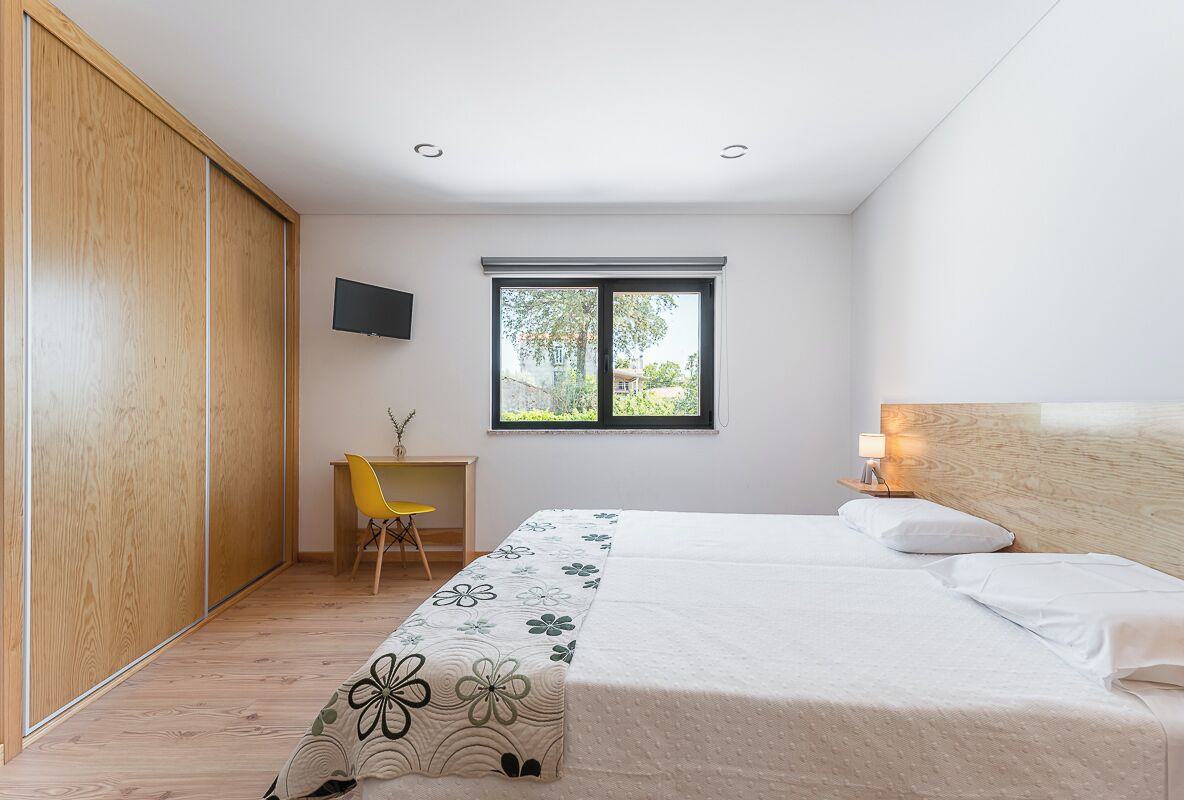 A bedroom with a bed, a desk, a chair, a TV, and a window.