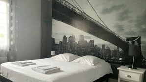 Une chambre d’hôtel moderne avec un grand lit, du linge de lit blanc, une table de chevet et une fresque représentant un paysage urbain sur le mur.