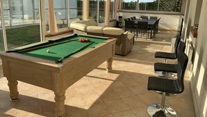 Une table de billard avec des billes, un canapé et un coin repas avec des chaises et une table, le tout dans une pièce lumineuse grâce à de grandes fenêtres.