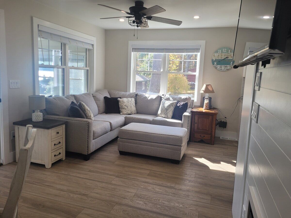 A living room with a sectional sofa, ottoman, wooden flooring, ceiling fan, and a window with blinds.