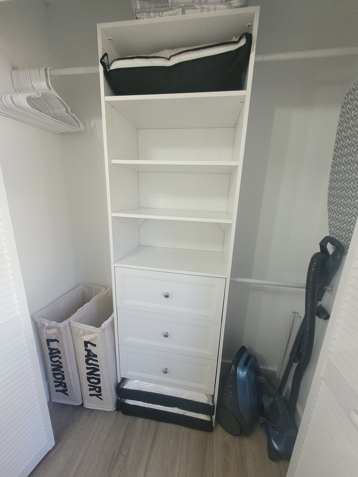 A white storage unit with multiple shelves and drawers, a vacuum cleaner, and two laundry bins.