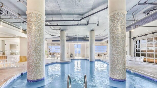 An indoor swimming pool with clear water, surrounded by columns with mosaic patterns, and a view of the sky through large windows.