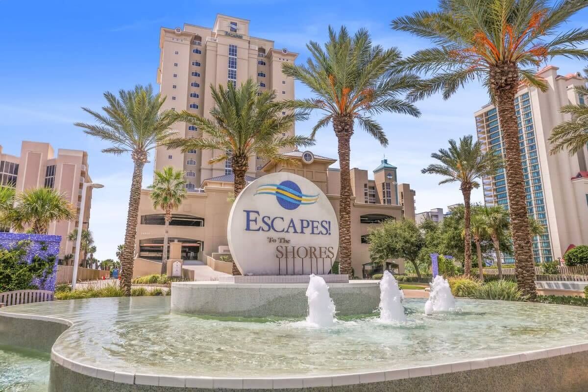 A hotel entrance with a sign reading 'Escapes! To The Shores,' palm trees, and a water feature.