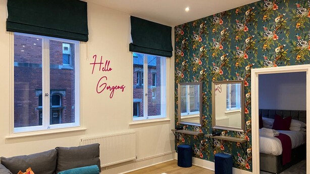 A modern living room with a grey sofa, a coffee table, and a bed with red pillows. A wall with a floral wallpaper and a 'Hello Gorgeous' sign is visible.