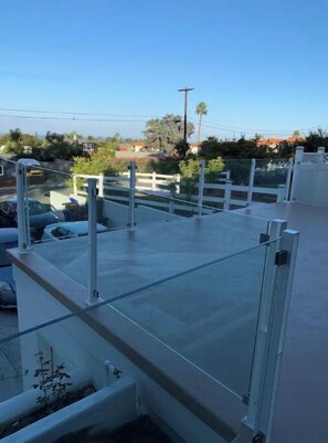 A rooftop terrace with a glass railing, a concrete floor, and a view of the surrounding neighborhood.