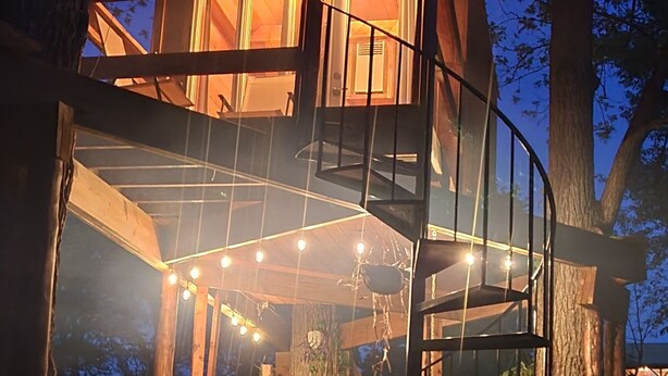 Ein hölzernes Baumhaus mit Wendeltreppe, umgeben von Bäumen und beleuchtet mit Lichterketten.