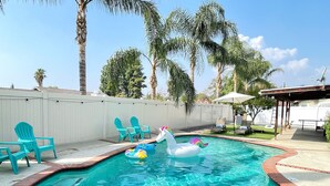 A backyard swimming pool with inflatable unicorn floats, surrounded by palm trees and a white fence. - Family 3BR POOL Home/ KING BED Suite/ BILLIARD TABLE/ Sleeps 9/near Yaamava' (Rialto)