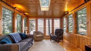 Un interior de cabaña acogedor, con un sofá, sillones y un ventanal que ofrece vistas al bosque.
