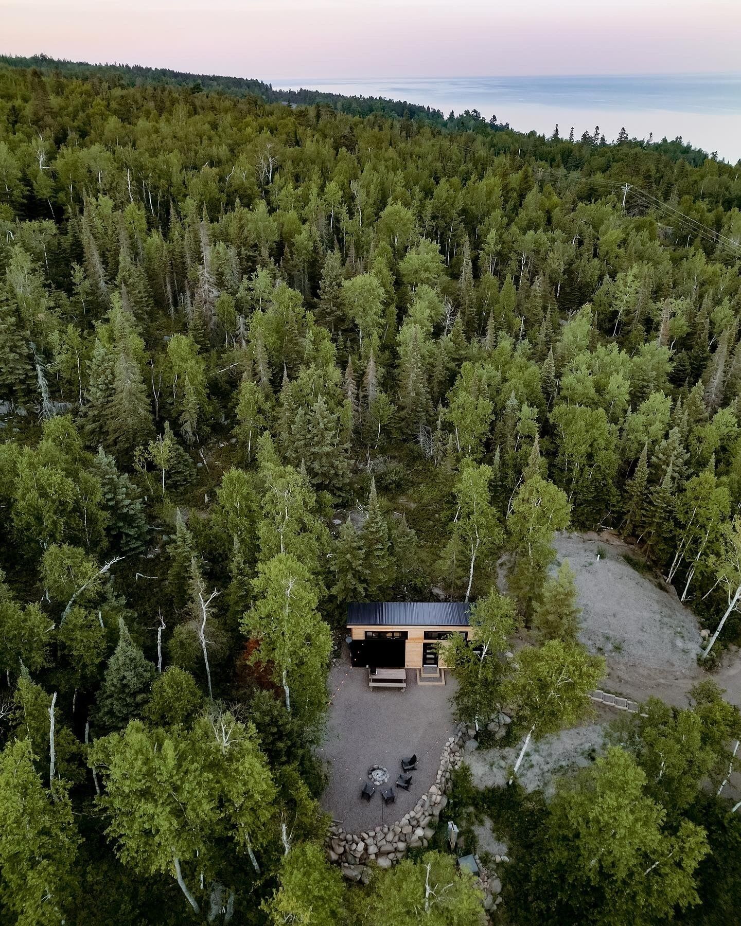 Una cabaña aislada rodeada de un denso bosque, con un área de grava y mobiliario exterior.