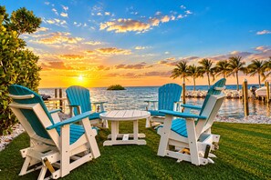 A sunset view with a table and four Adirondack chairs on a grassy area by the water.