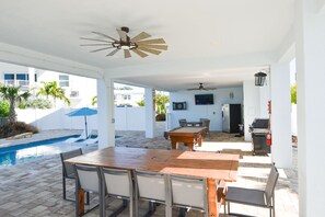 Une terrasse extérieure avec une piscine, une table et des chaises en bois, un ventilateur de plafond et un barbecue.