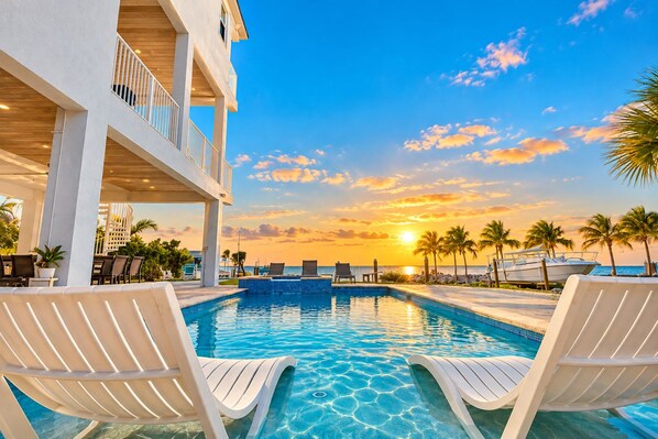 A pool area with lounge chairs, a view of the sunset, and a building with a balcony.