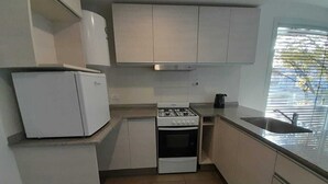 Een kleine keuken met witte kastjes, een ingebouwd gasfornuis, een kleine koelkast en een raam met jaloezieën.