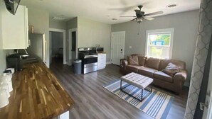 Una cocina moderna con encimera de madera, horno empotrado y microondas. En la zona de estar, hay un sofá color beige y un pequeño otoman.
