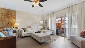 A spacious living room with a sofa, armchair, and ottoman, a ceiling fan, and a large window with curtains. - Location, location, location! Steps to the pool and just a 5-minute walk to the beach 🏖️💦 (Miramar Beach)