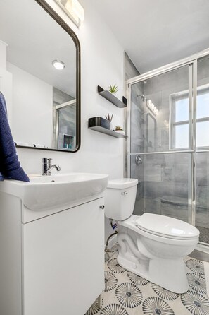 A modern bathroom with a white sink, toilet, and shower area. There are two wall-mounted shelves with plants and a large mirror above the sink. - Best Little Bunkhouse in TX! Walk to FIFA WORLD CUP (Arlington)