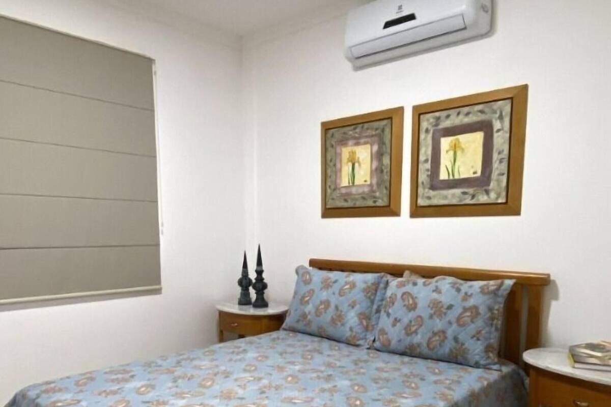 A bedroom with a bed, an air conditioner, two framed pictures, a bedside table, and a window with blinds.