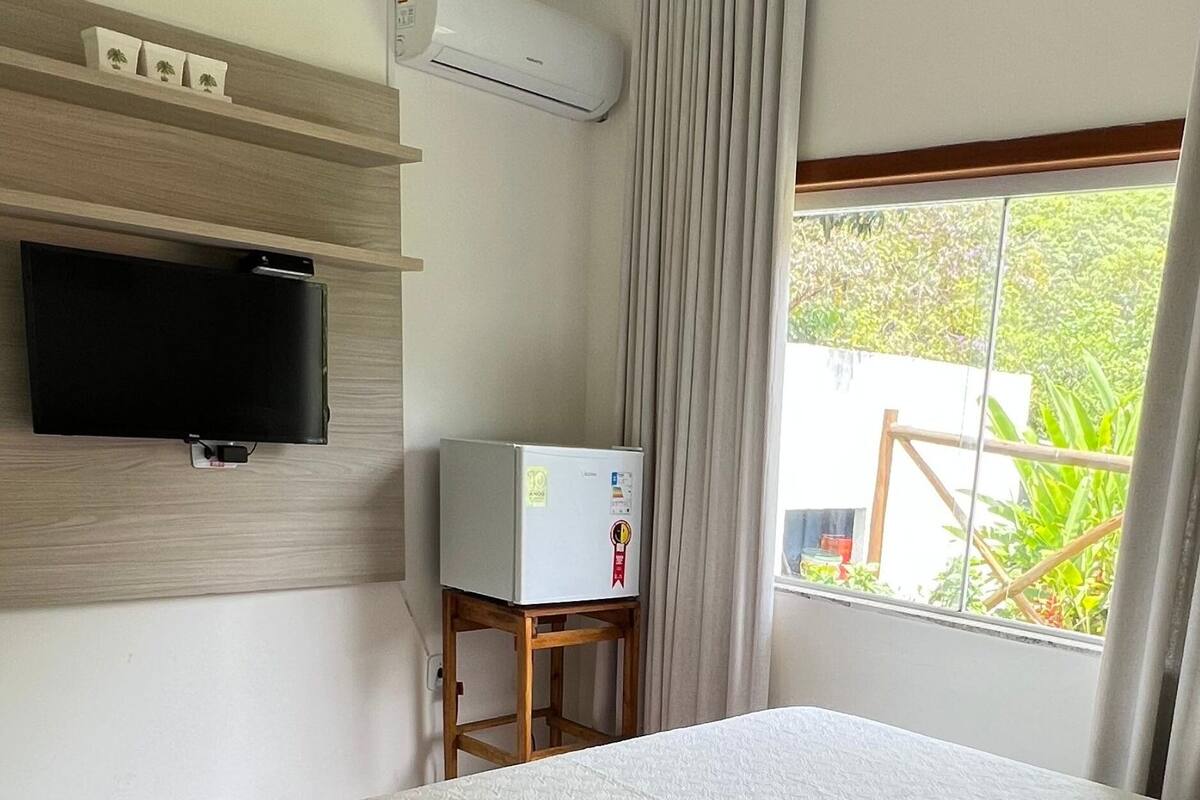 A hotel room with a bed, a television mounted on the wall, a small refrigerator on a wooden table, and a window with a view of greenery.