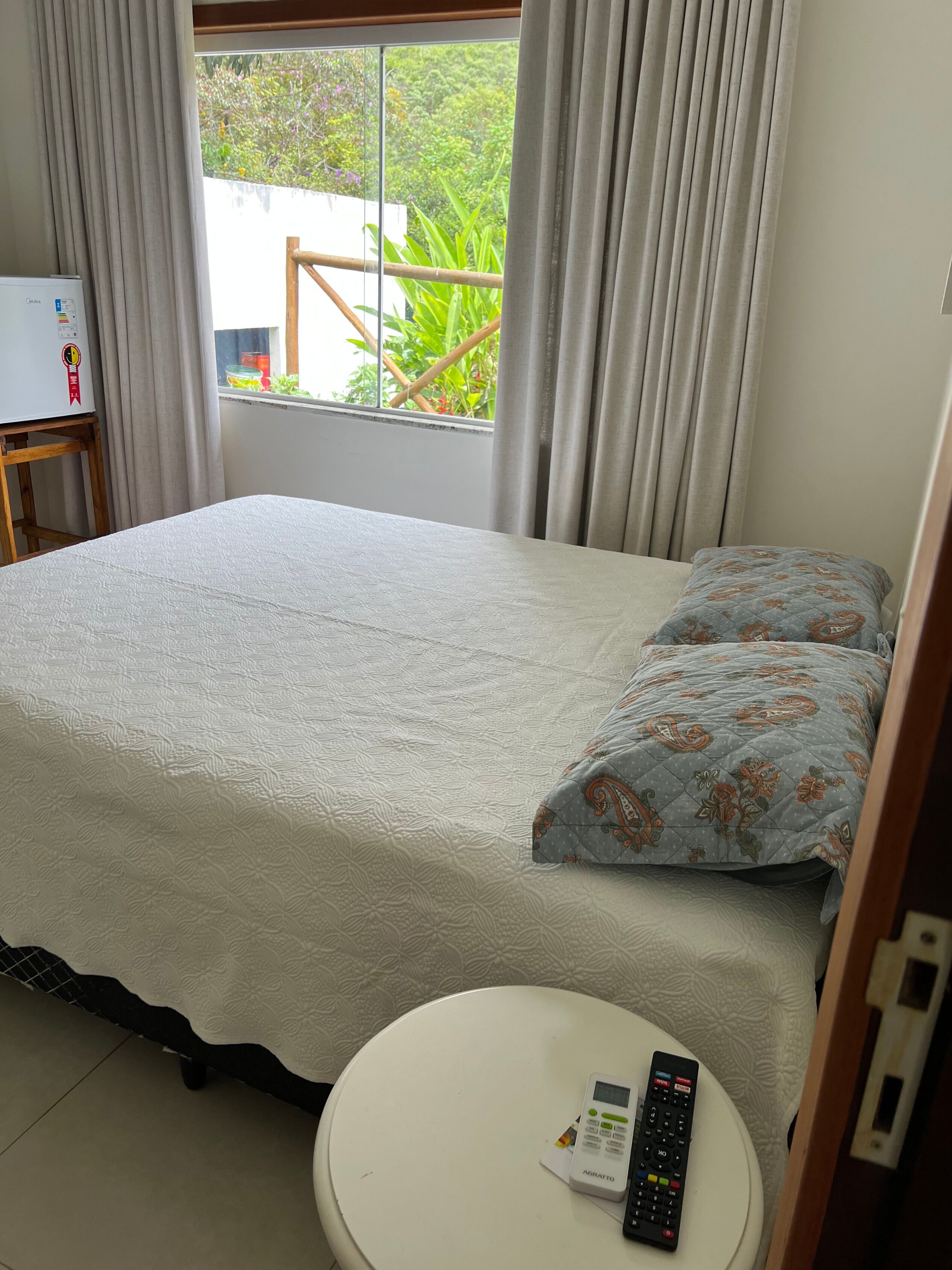 A bedroom with a bed, a bedside table, a remote control, and a window with curtains.