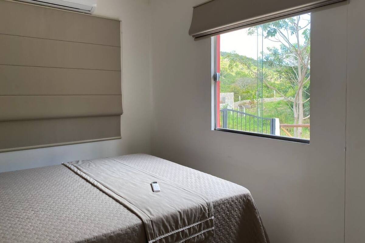 A room with a bed, a window with a view of greenery, and an air conditioning unit.