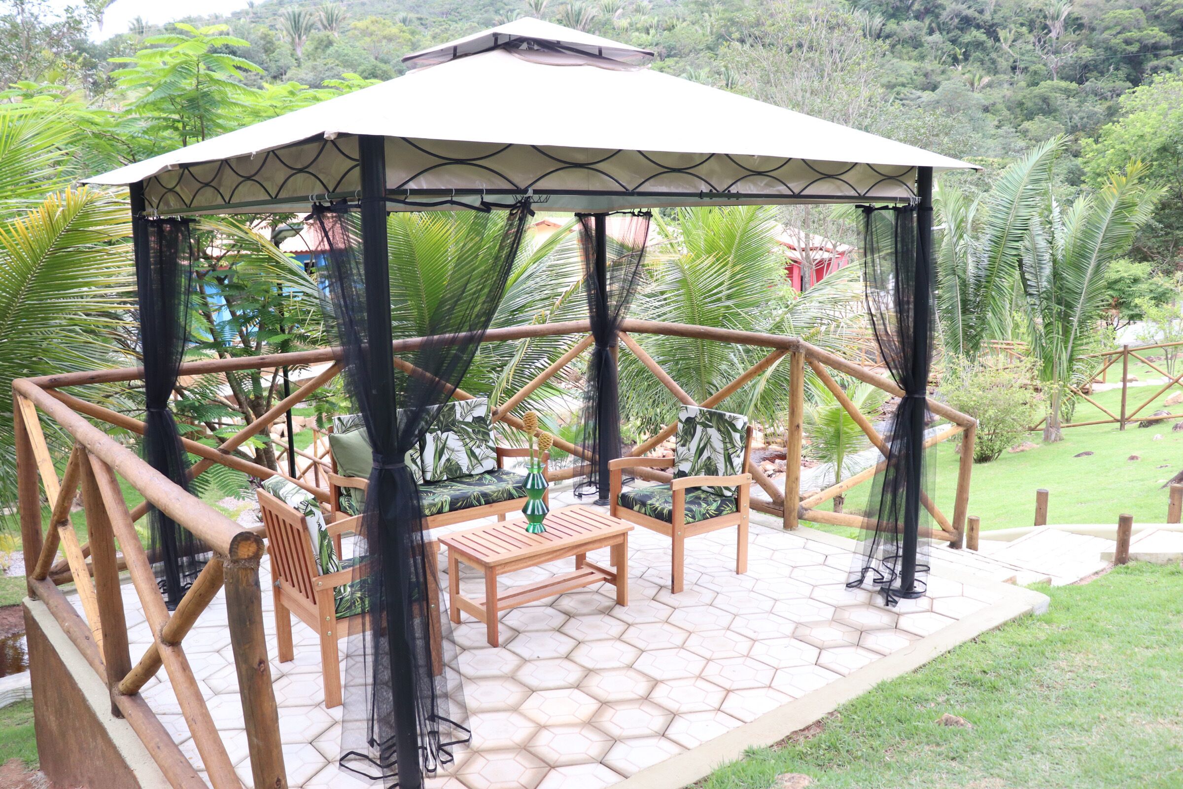 Um gazebo de madeira com piso hexagonal em mosaico, cercado por vegetação exuberante e um pequeno lago.