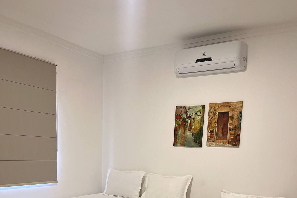 A hotel room with two single beds, a wooden nightstand, an air conditioning unit, and two wall-mounted paintings.