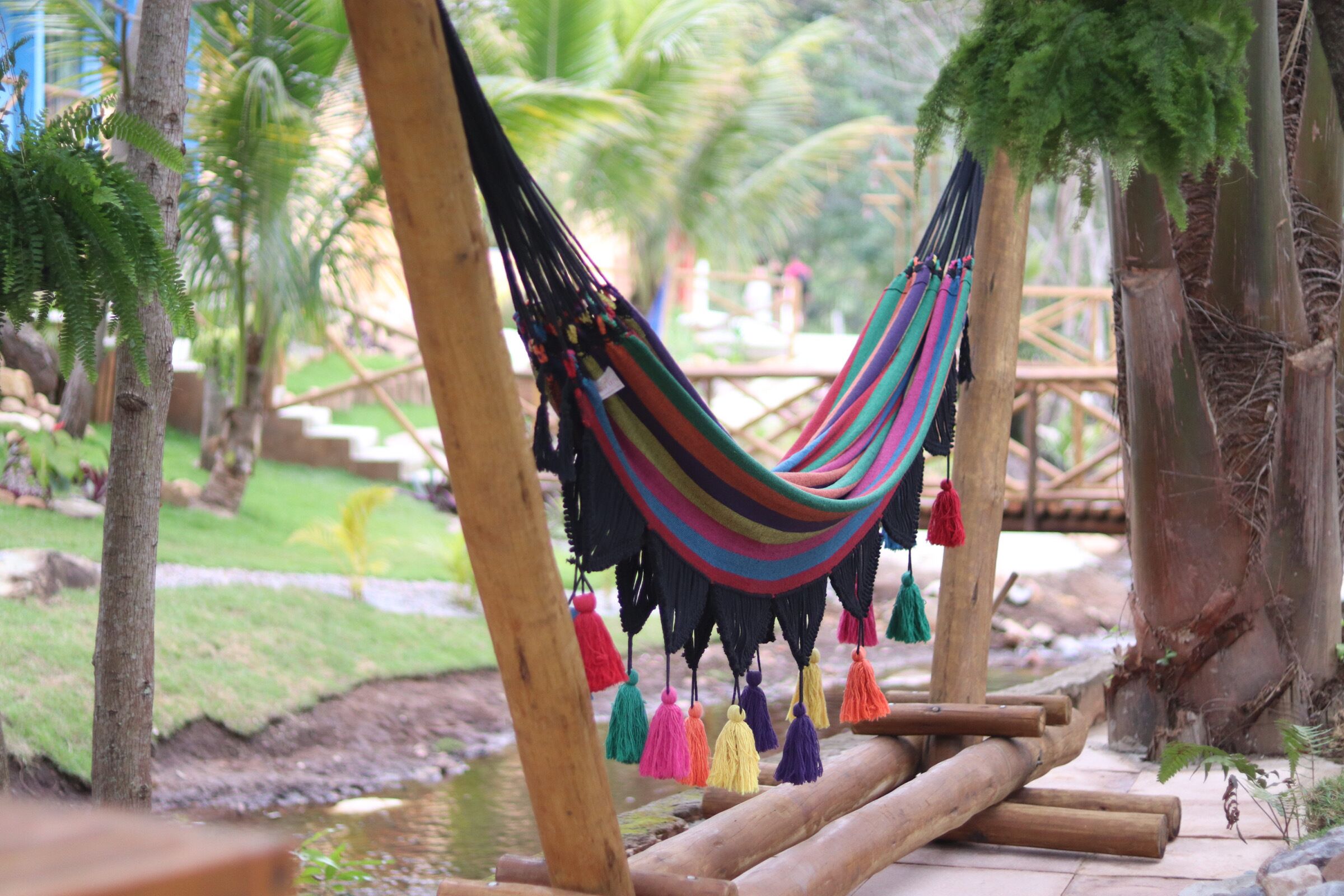 A hammock suspended between wooden posts in a garden with palm trees and a wooden bridge.