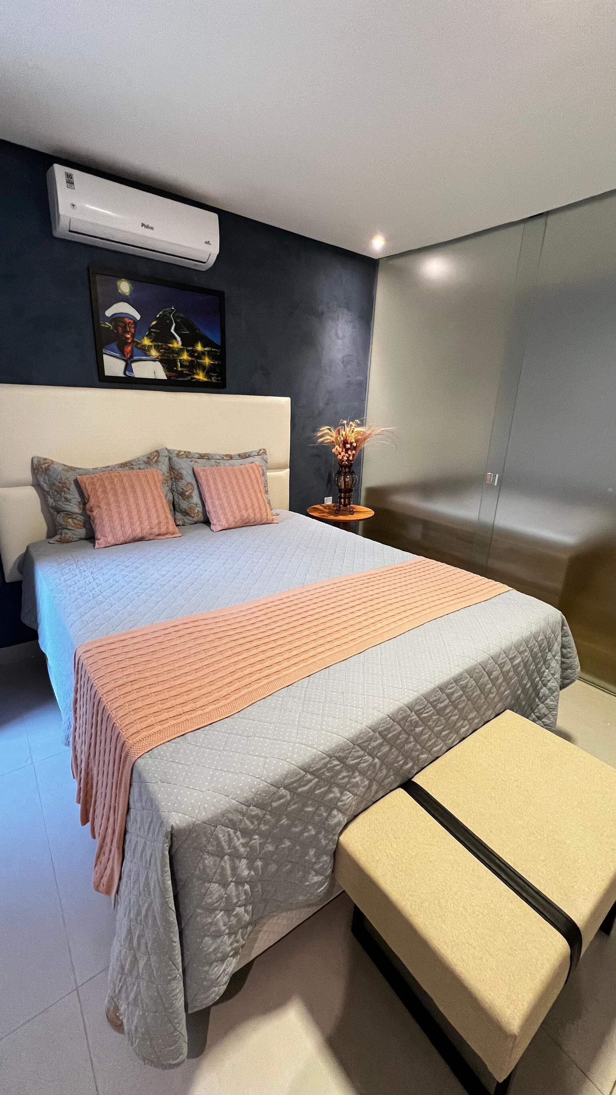 A modern bedroom with a large bed, a bedside table with a vase, and a wall-mounted air conditioner.