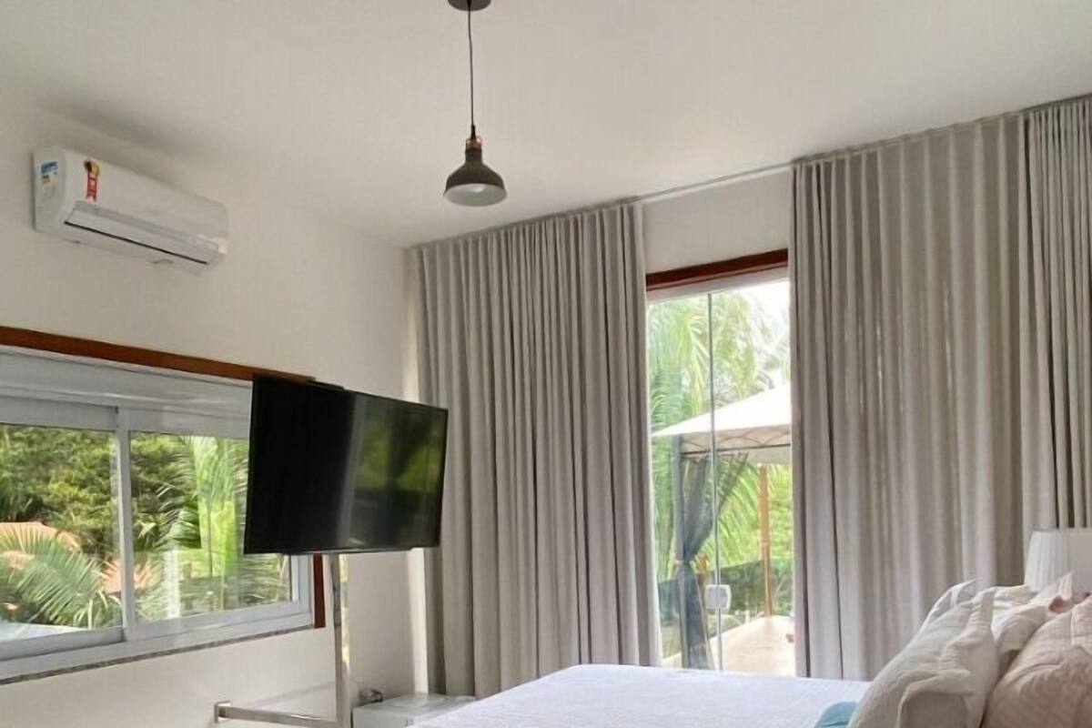 A bedroom with a large bed, a flat-screen TV, an air conditioning unit, and a window with curtains.