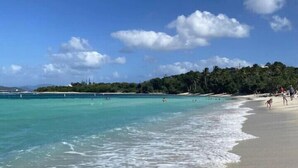 Ein Strand mit klarem türkisblauem Wasser, einem sandigen Ufer und einer entfernten Hügelkette mit viel Grün.
