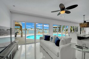 Un salon moderne avec un canapé blanc, une table basse en verre et un ventilateur de plafond. De grandes fenêtres offrent une vue sur une piscine et du mobilier d’extérieur.