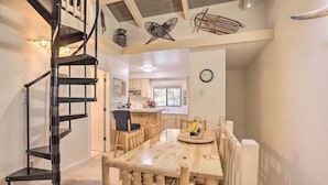 Un espace repas avec une table et des chaises en bois, un escalier en colimaçon noir et, au fond, une cuisine.