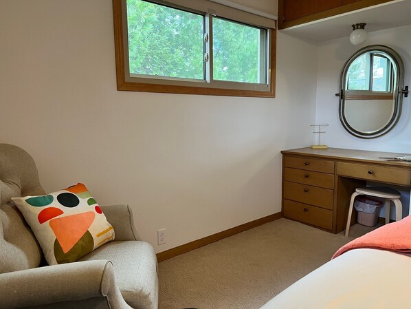 Un dormitorio con cama, una silla, un escritorio, un espejo y ventana con vista a árboles.