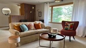 A living room with a beige sofa, a red armchair, a round coffee table, and a small side table. There is a window with curtains, a mirror on the wall, and a wooden slatted partition. - Beautifully styled, Mid Century Modern, family friendly Lake Mills House! (Lake Mills)